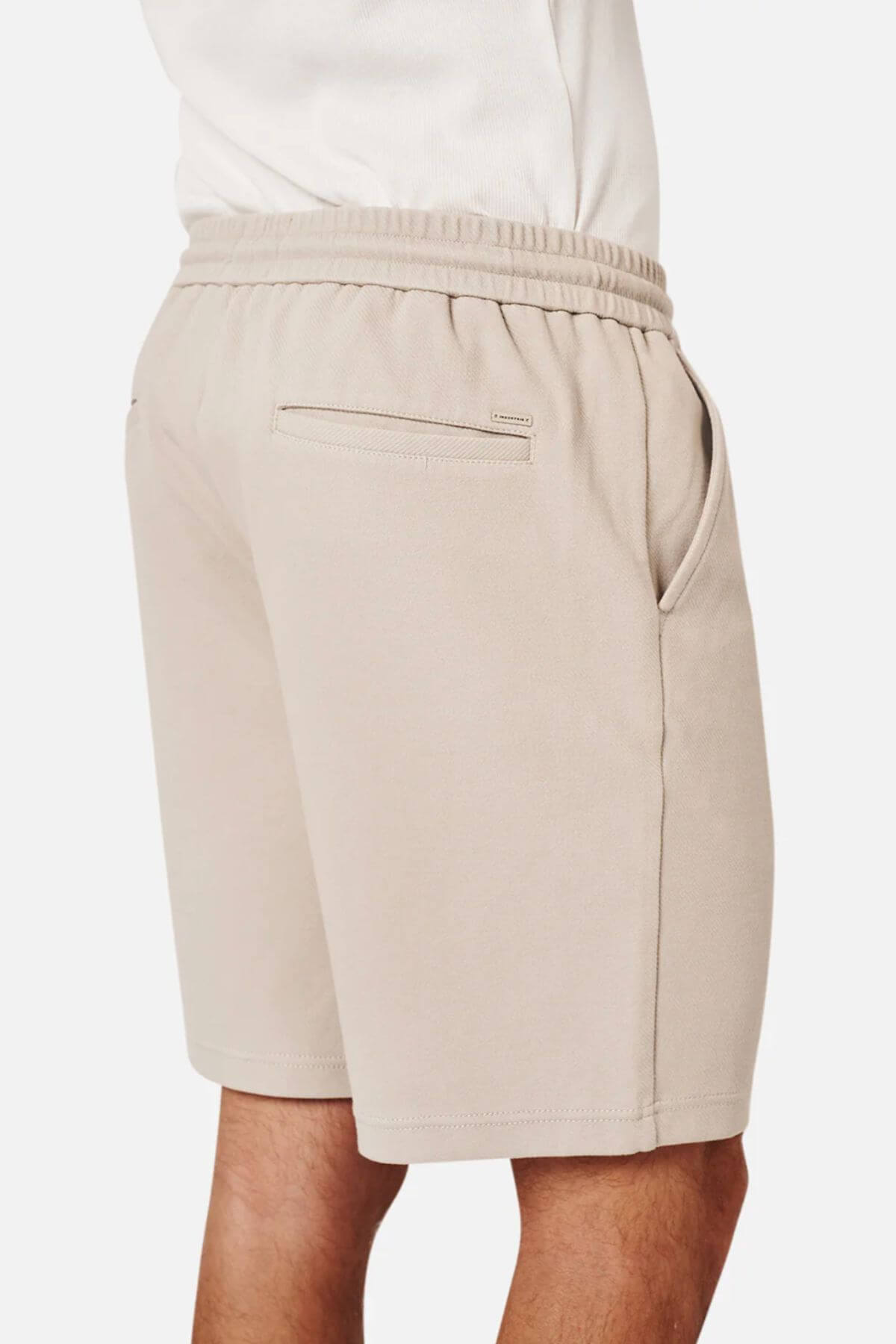 A person wears Industrie The Broadway Short in light beige, featuring an elastic waistband, side pockets, relaxed fit, and a small zippered back pocket, shown from the back and side.