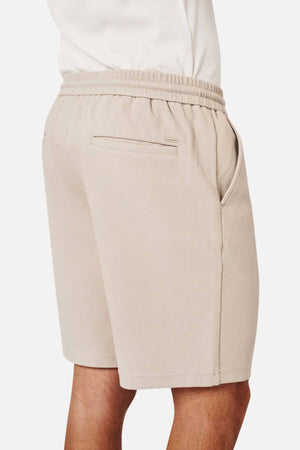 A person wears Industrie The Broadway Short in light beige, featuring an elastic waistband, side pockets, relaxed fit, and a small zippered back pocket, shown from the back and side.