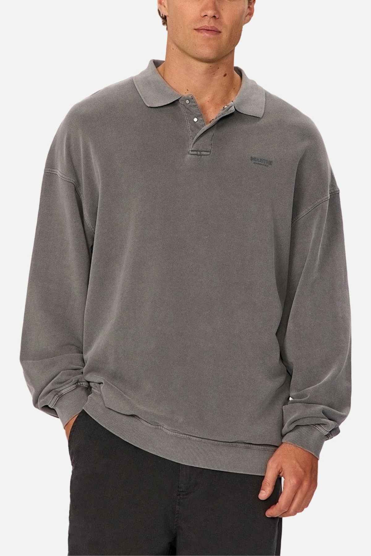 A person models the Industrie The Burbank Polo Track, a loose-fitting, long-sleeve gray collared shirt with buttons, paired with dark pants. Styled simply against a white background, one hand rests in their pocket.