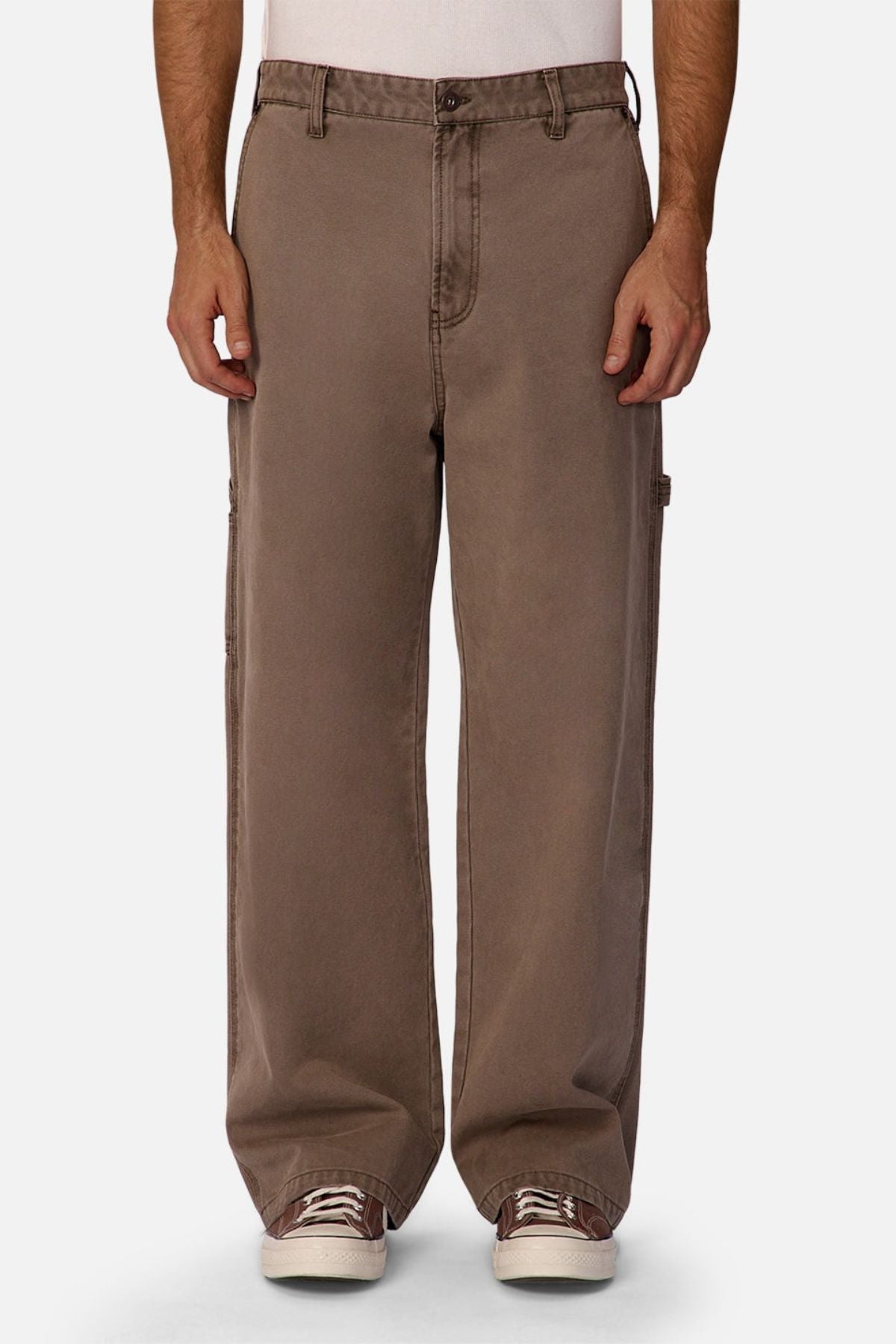 A person wears Industrie The Carolina Carpenter Pant—loose-fitting, durable cotton pants in brown with visible side pockets and stitching—paired with white sneakers, standing against a plain white background.