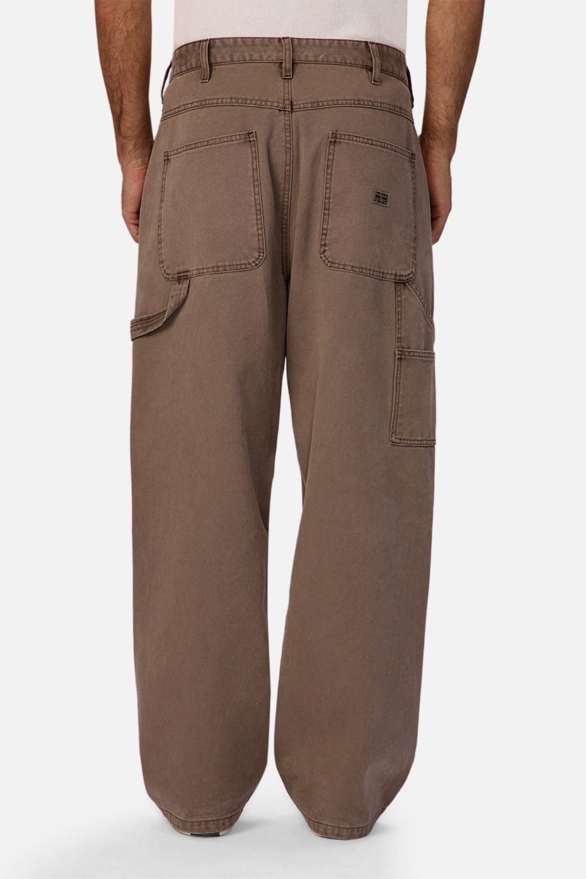 A person wearing Industrie The Carolina Carpenter Pant in loose-fitting brown cotton, viewed from the back, showcasing large square back pockets, a side pocket, and visible stitching.
