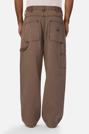 A person wearing Industrie The Carolina Carpenter Pant in loose-fitting brown cotton, viewed from the back, showcasing large square back pockets, a side pocket, and visible stitching.