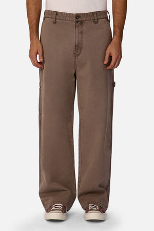A person wears Industrie The Carolina Carpenter Pant—loose-fitting, durable cotton pants in brown with visible side pockets and stitching—paired with white sneakers, standing against a plain white background.