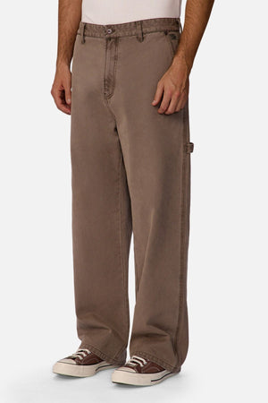 A person wearing the Industrie The Carolina Carpenter Pant in loose-fitting brown cotton, paired with a white shirt and brown sneakers, shown from shoulders to feet against a plain white background.