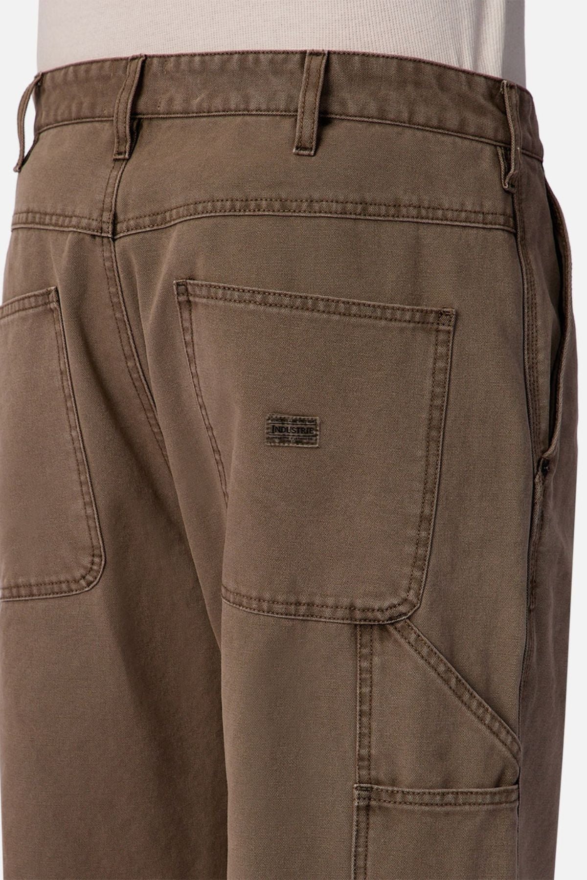 Close-up of a person wearing Industrie The Carolina Carpenter Pant in brown, featuring visible back pockets, detailed stitching, and a small rectangular label on one pocket. Durable cotton fabric is styled with a tucked-in light-colored shirt.