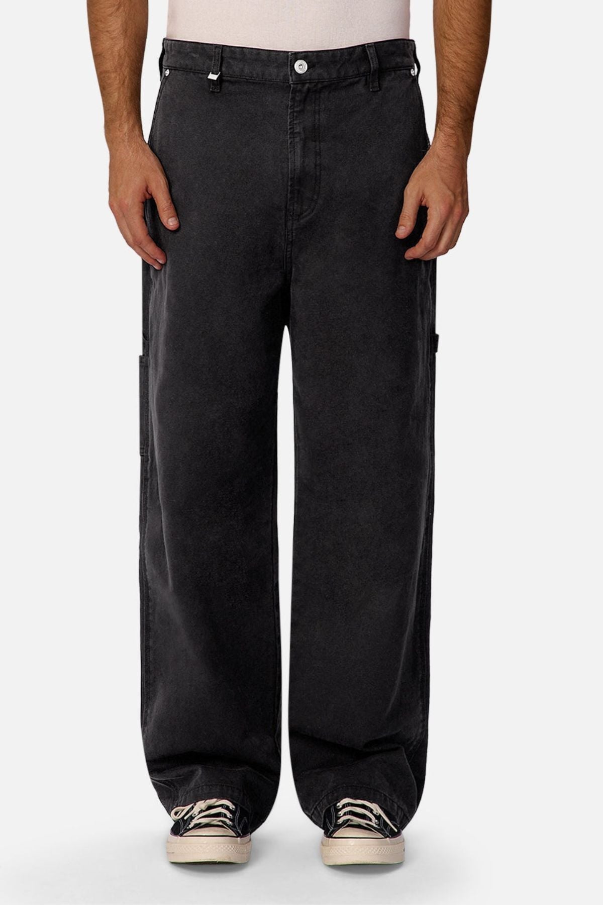 A person wearing Industrie The Carolina Carpenter Pant in black with side pockets, a white shirt tucked in, and white sneakers, standing against a plain light background.