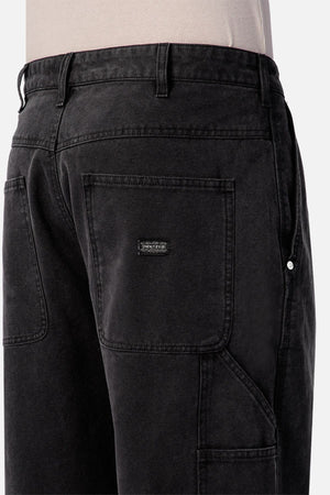 A close-up of someone wearing Industrie The Carolina Carpenter Pant in black denim cotton, showcasing large square pockets, visible belt loops, and a small rectangular black patch on one pocket.