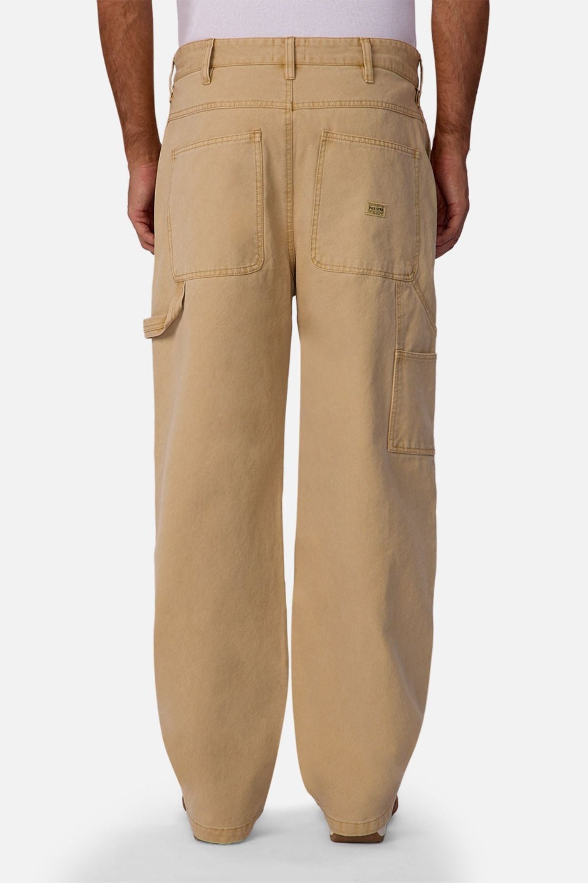 A person wearing Industrie The Carolina Carpenter Pant, featuring loose-fitting, durable cotton fabric with large back pockets and a side tool pocket, shown from the back against a plain white background.
