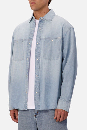 A person wears the Industrie The Chambray Nebraska Long Sleeve Shirt—light blue with two chest pockets and silver buttons—over a white t-shirt, paired with matching light blue denim jeans against a plain white background, reflecting classic workwear style.