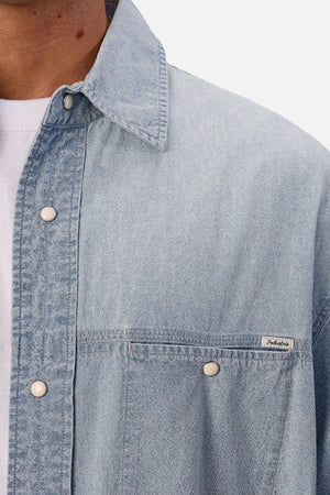 Close-up of a person wearing the Industrie The Chambray Nebraska Long Sleeve Shirt in light blue over a white tee, with white snap buttons and a chest pocket label. Only the lower face and upper torso evoke classic men's workwear style.