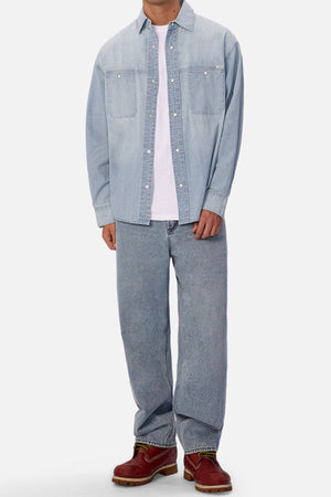 Wearing the Industrie The Chambray Nebraska Long Sleeve Shirt over a white tee, loose light blue jeans, and brown leather boots, a person stands against a plain light background. Classic men's workwear style; head not visible.