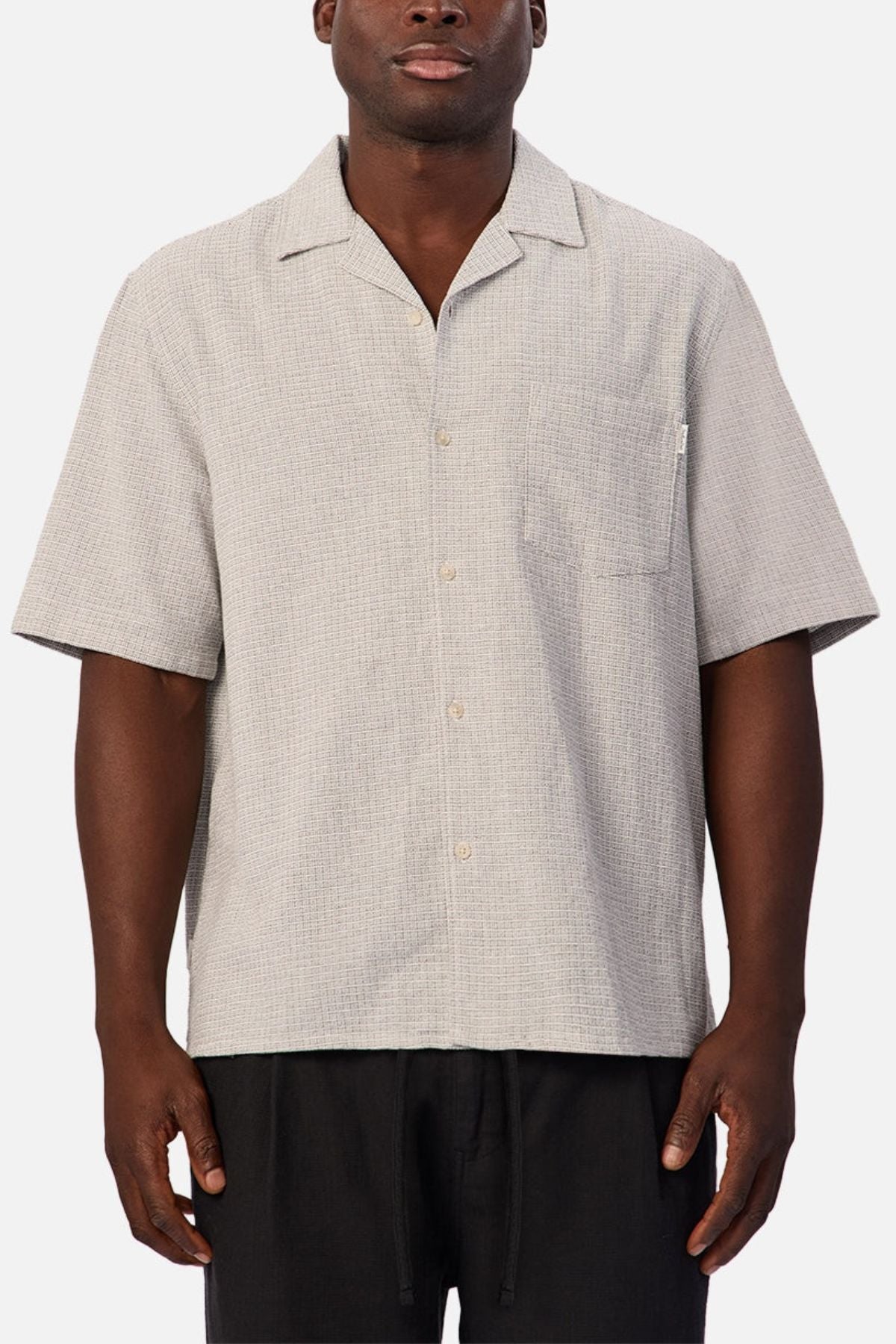 A person is wearing the Industrie The Check Marbella Short Sleeve Shirt, a light grey short sleeve casual shirt with a chest pocket, paired with black pants, standing against a plain background.