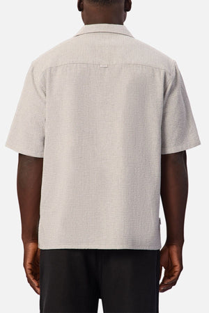 A person stands facing away, wearing the Industrie The Check Marbella Short Sleeve Shirt in light gray textured fabric paired with black pants. The casual shirt features a relaxed fit, a straight hem, and a seam across the upper back. The background is plain.