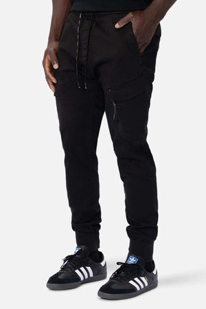 A person is wearing black Industrie The Defender Combat Pants with a drawstring waistband and side pockets, ideal for an active lifestyle. They're also sporting black Adidas sneakers with white stripes, standing confidently against a plain white background.