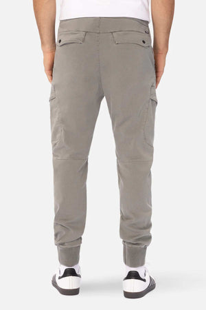 A person wearing Industrie The Defender Combat Pant in gray cargo jogger style and white sneakers with black accents, standing with their back turned. These active lifestyle pants feature buttoned back pockets, elastic cuffs at the ankles, and offer durable comfort. The background is plain white.