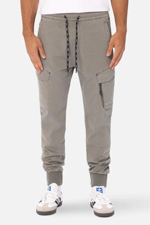A person is wearing Industrie The Defender Combat Pant in light gray with an elastic waistband and drawstring closure. They have their hands in the side and cargo pockets of the pants. The person is also sporting white sneakers with blue and gray accents, all set against a plain white background.