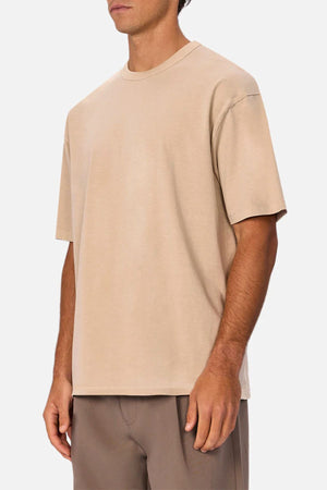 A man wears an oversized beige Industrie The Del Sur Tee made from heavyweight fabric with light brown pants, standing against a plain white background. His face is partially out of frame.
