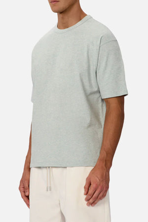 A man wears an oversized Industrie The Del Sur Tee in light gray heavyweight fabric with cream drawstring pants, standing against a plain light background. His face is partially out of frame and his arms are relaxed at his sides.