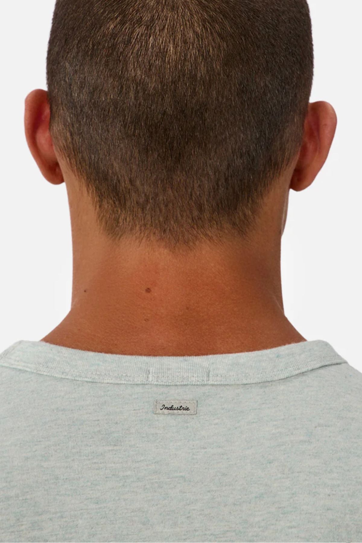 A close-up of the back of a person's head shows short brown hair and a light gray Industrie The Del Sur Tee with a small "Industrie" label on the collar. The tee is made from heavyweight fabric, set against a plain white background.