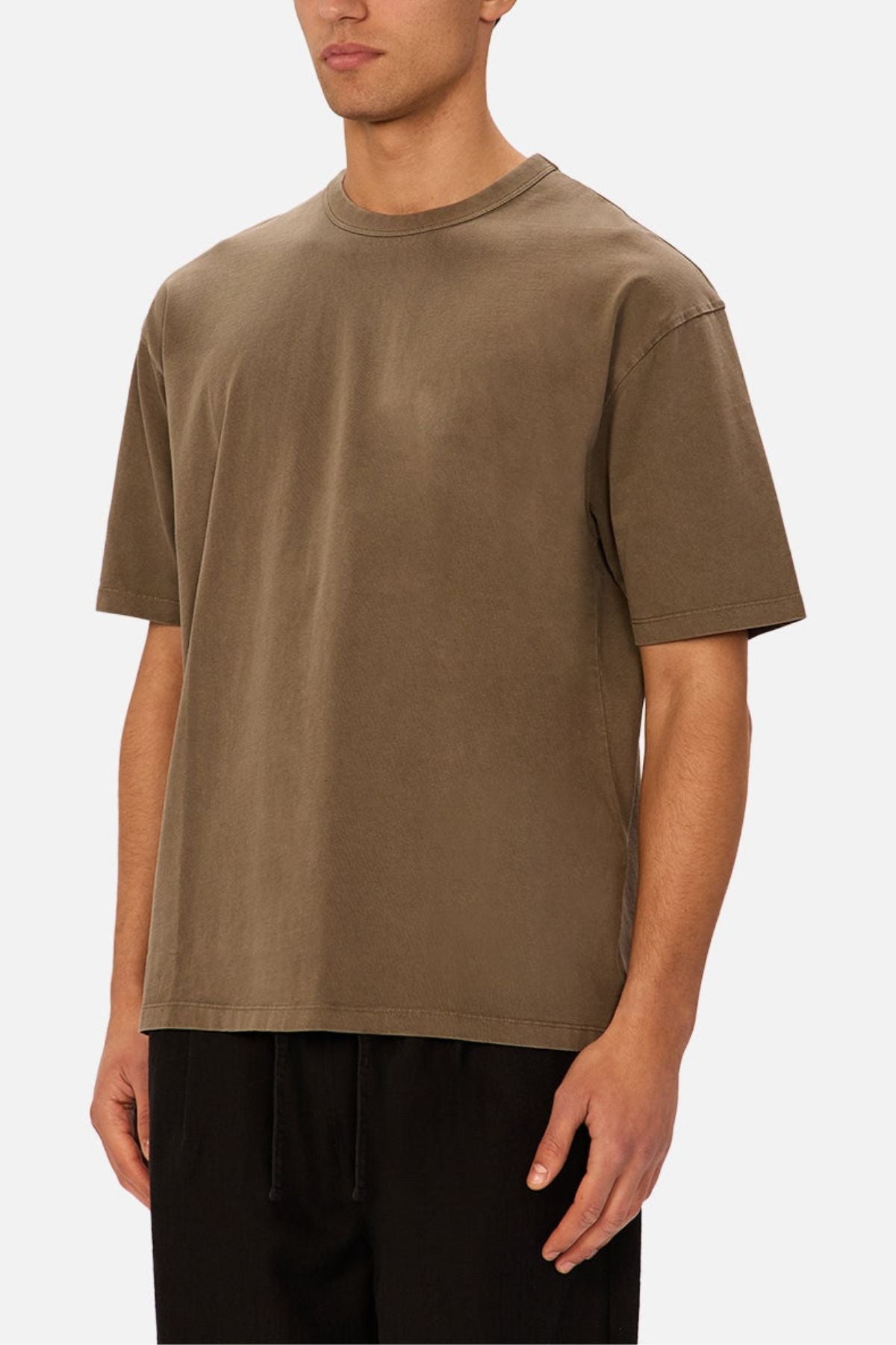 A person wearing the Industrie The Del Sur Tee in brown—a loose-fitting, short-sleeve shirt crafted from heavyweight cotton—paired with black pants, stands against a plain light background. The face is partially visible, and the oversized fit of the outfit appears casual and comfortable.