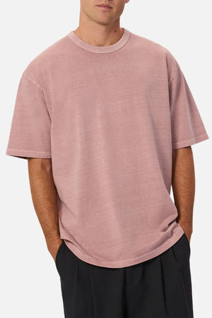 Someone stands with hands in their pockets against a plain background, wearing the Industrie The Del Sur Tee—an oversized fit t-shirt in light pink crafted from heavyweight fabric.