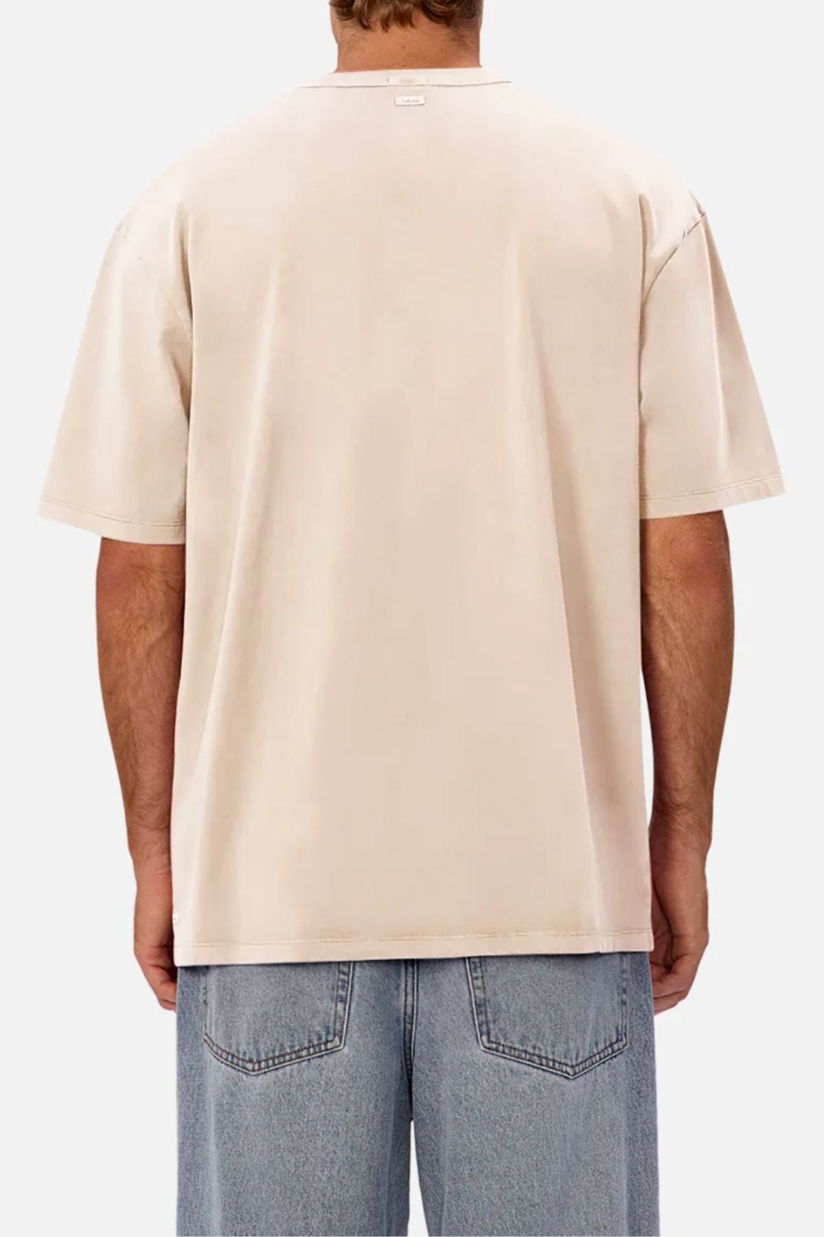 A person is seen from behind against a white background, wearing a loose, light beige Industrie The Del Sur Tee and blue jeans—the oversized t-shirt offers a relaxed and comfortable fit.