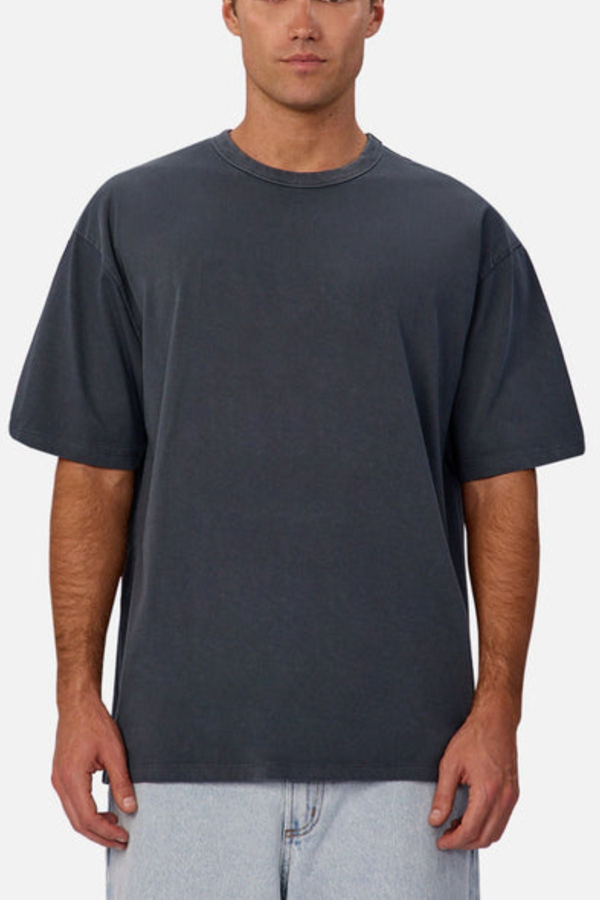 A man in an oversized Industrie The Del Sur Tee, short-sleeved and dark gray, made from heavyweight cotton, with light blue jeans stands against a plain light background. His expression is neutral and his face is partially visible.