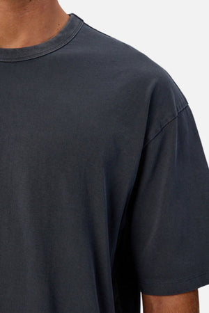 A close-up shows a person wearing the Industrie The Del Sur Tee, an oversized, plain dark gray heavyweight cotton shirt; only their upper torso and part of their neck are visible against a light background.