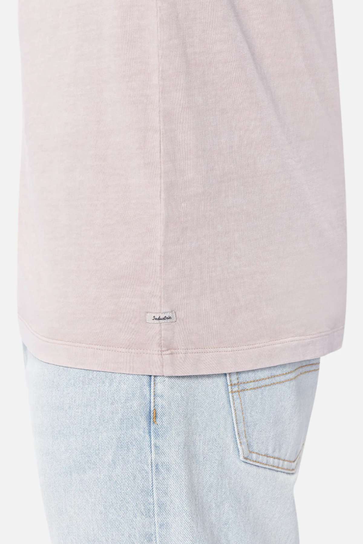A person wearing a light pink Industrie The Del Sur Tee and light blue jeans. The focus is on the lower part of the 100% cotton t-shirt, which has a small tag with text on the side seam, and the upper part of the jeans, showing a back pocket with visible stitching.