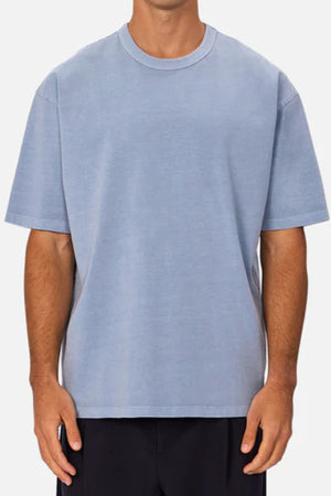A person, head not visible, wears an oversized Industrie The Del Sur Tee in light blue with black pants, standing against a plain light background.
