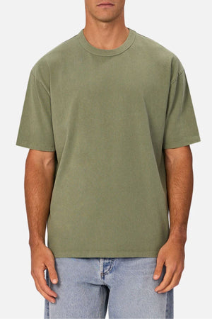 Someone wearing an oversized olive green Industrie The Del Sur Tee, crafted from heavyweight fabric, paired with light blue jeans and standing against a plain background.