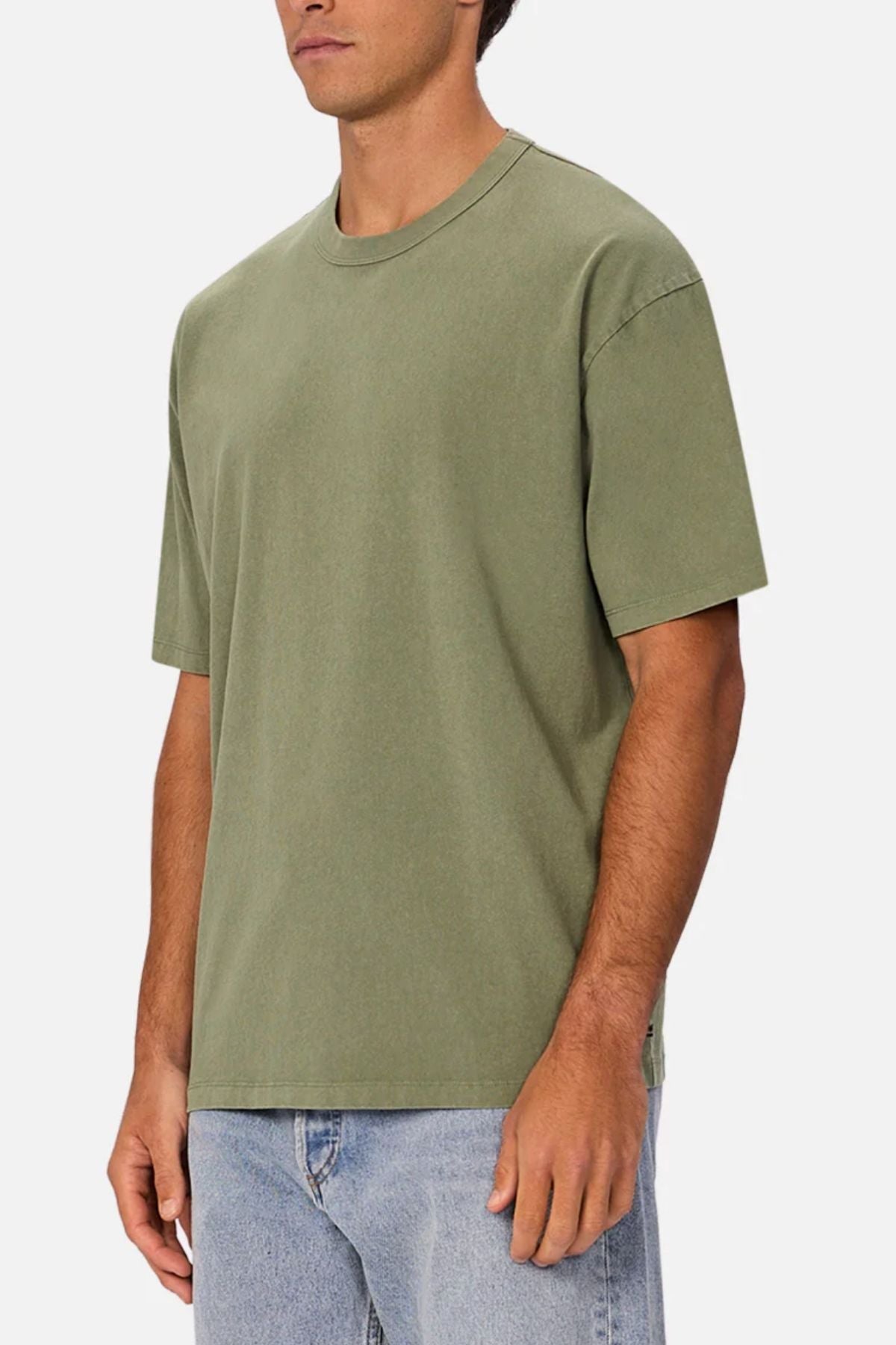 A man wearing the Industrie The Del Sur Tee—an oversized, heavyweight olive green t-shirt—and light blue jeans stands against a plain white background with his face partially out of frame.