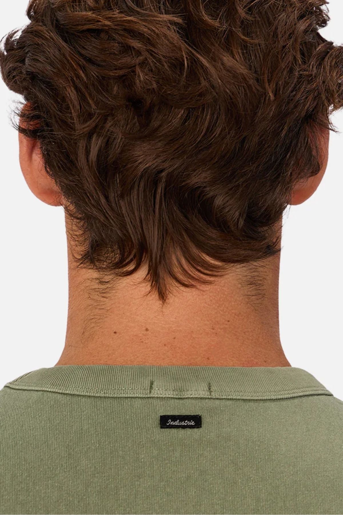 The image shows the back of a person with wavy brown hair wearing an oversized sage green Industrie The Del Sur Tee made of heavyweight fabric, featuring a small black "Industrie" label on the collar.