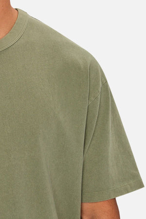 Close-up of a person wearing the Industrie The Del Sur Tee, an oversized olive green heavyweight shirt. Only the upper torso and part of one arm are shown against a plain light background.
