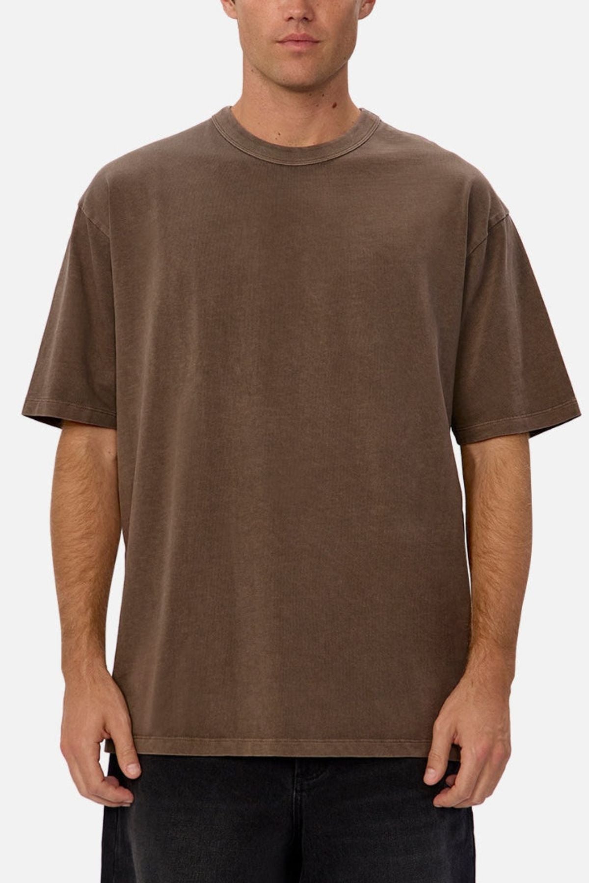 An individual stands facing forward against a plain background, wearing the Industrie The Del Sur Tee in brown—a loose-fitting, heavyweight cotton t-shirt with a simple crew neckline—paired with dark pants and featuring no visible logos or designs.