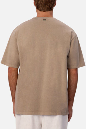 A person is seen from behind wearing an oversized Industrie The Del Sur Tee in beige with short sleeves and plain back, paired with light-colored pants against a simple light background.