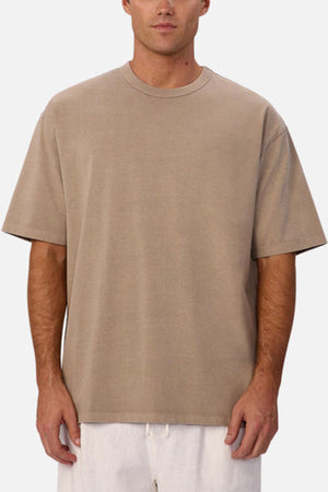 A man wears an oversized Industrie The Del Sur Tee in beige with white pants, standing against a plain white background. His face is partially visible.