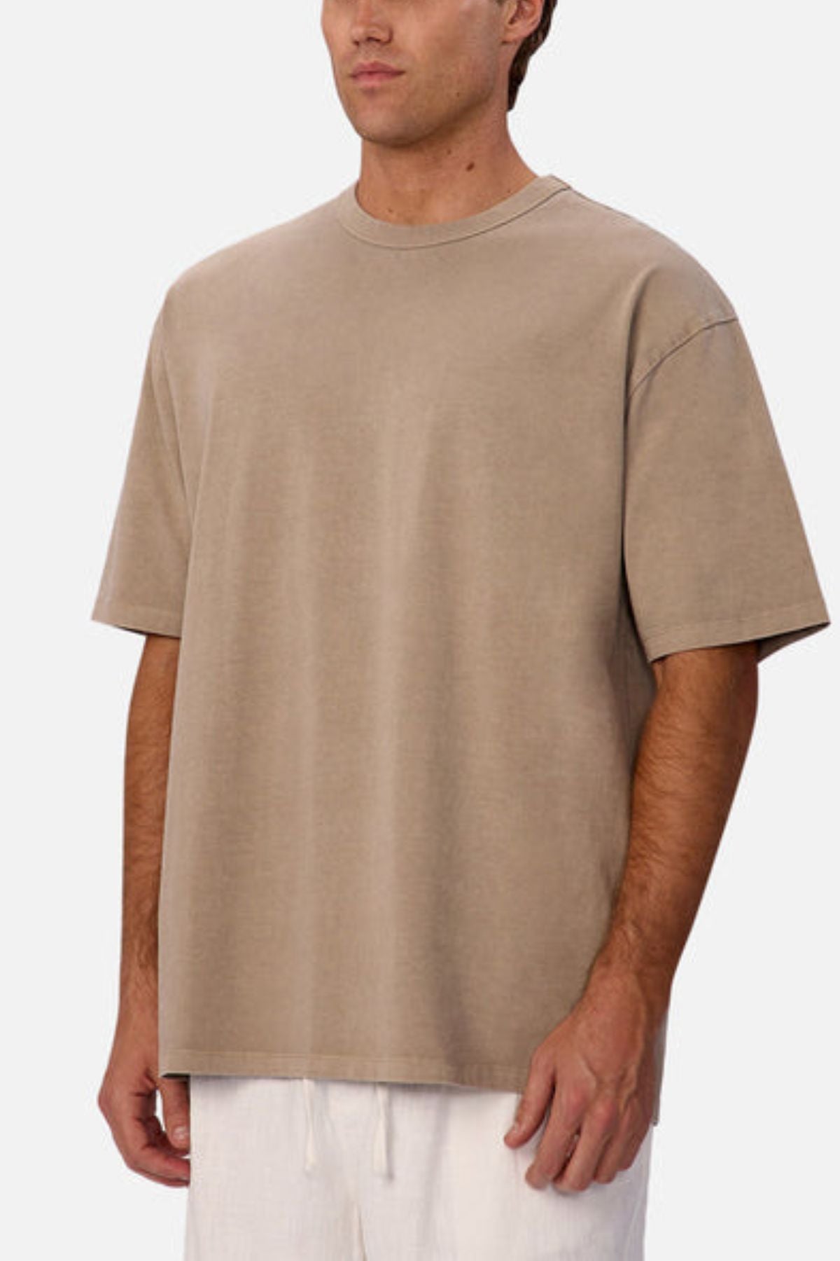 A man stands against a plain white background wearing the Industrie The Del Sur Tee in beige and white pants; his face is partially visible, and his arms are relaxed at his sides.