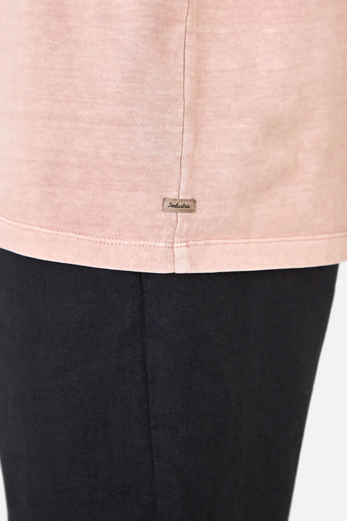Close-up of a person in Industrie The Del Sur Tee, a light pink oversized fit, 100% cotton shirt with a small metallic tag by the hem, paired with dark pants against a minimal background.