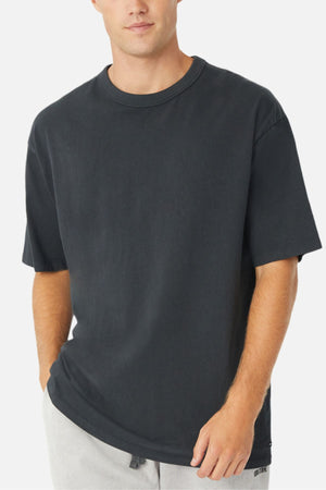 Wearing the Industrie The Del Sur Tee, a plain oversized 100% cotton black T-shirt, and light gray pants, a person stands against a white background with one hand in their pocket.