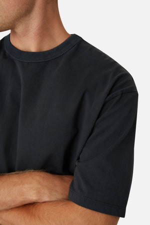 A person with light skin and short hair is shown from the shoulders up, wearing an oversized black Industrie The Del Sur Tee made of 100% cotton with arms crossed. The background is light gray, and their face is partially out of frame.