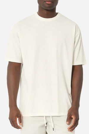 A person wearing an oversized Industrie The Del Sur Tee, made from 100% cotton, stands against a gray background. The off-white T-shirt features short sleeves and a loose fit, paired with light-colored pants, highlighting the simplicity of the outfit.