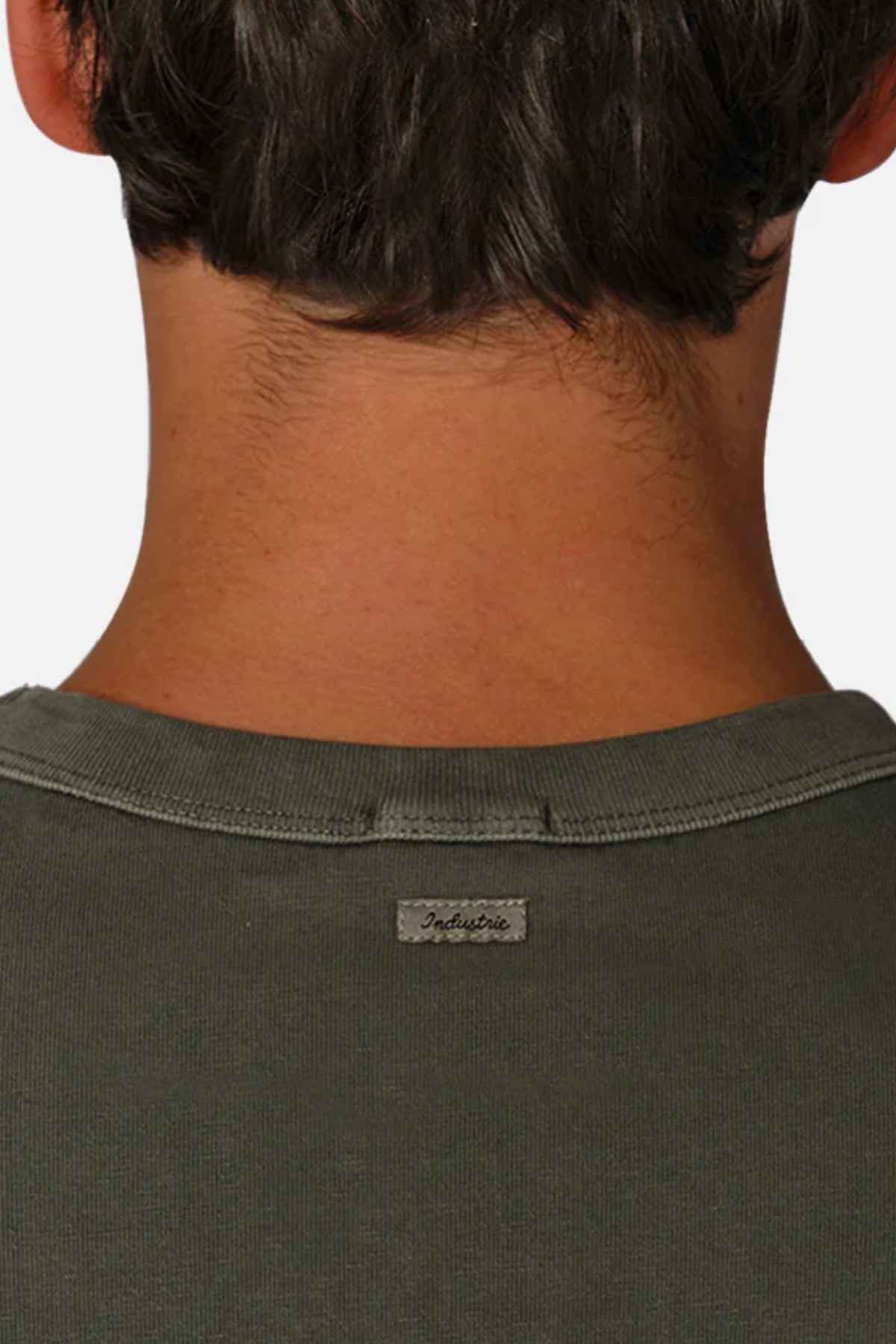 The image shows the back of a person's neck and head with dark hair, wearing the Industrie The Del Sur Tee in an oversized fit and dark green color. A small tag with text is visible on the collar, and the shirt appears to be made from soft fabric that might be 100% cotton. The background is plain and light-colored.