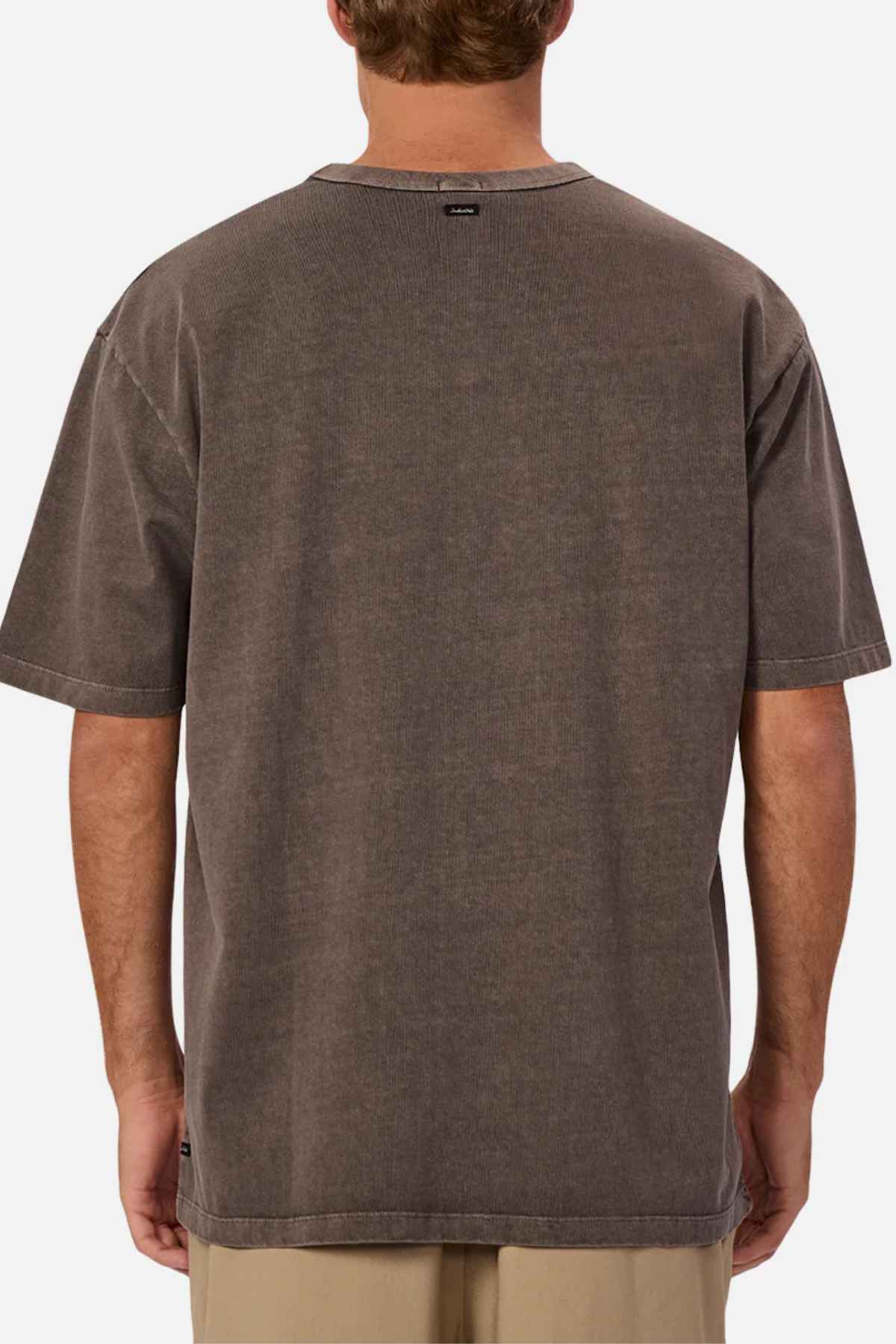 The person is showcasing an oversized fit, 100% cotton brown T-shirt—Industrie The Del Sur Tee—with a subtle tag at the neck on the back. Complemented by light-colored pants, they stand facing away from the camera, embodying a relaxed and stylish vibe.