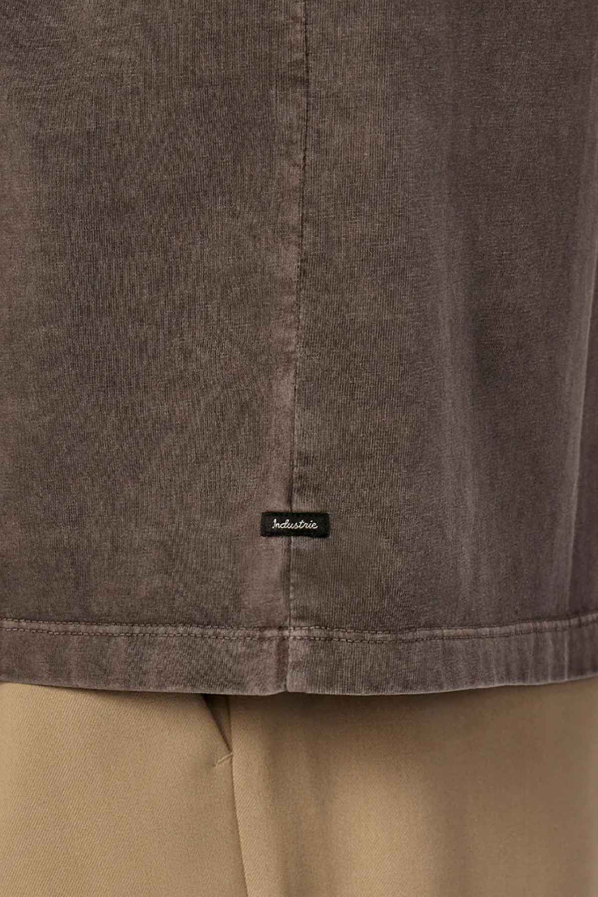 Close-up of the hem of The Del Sur Tee by Industrie, a brown oversized fit shirt made from 100% cotton, with a small black tag displaying a white logo. The tee is styled with beige pants.