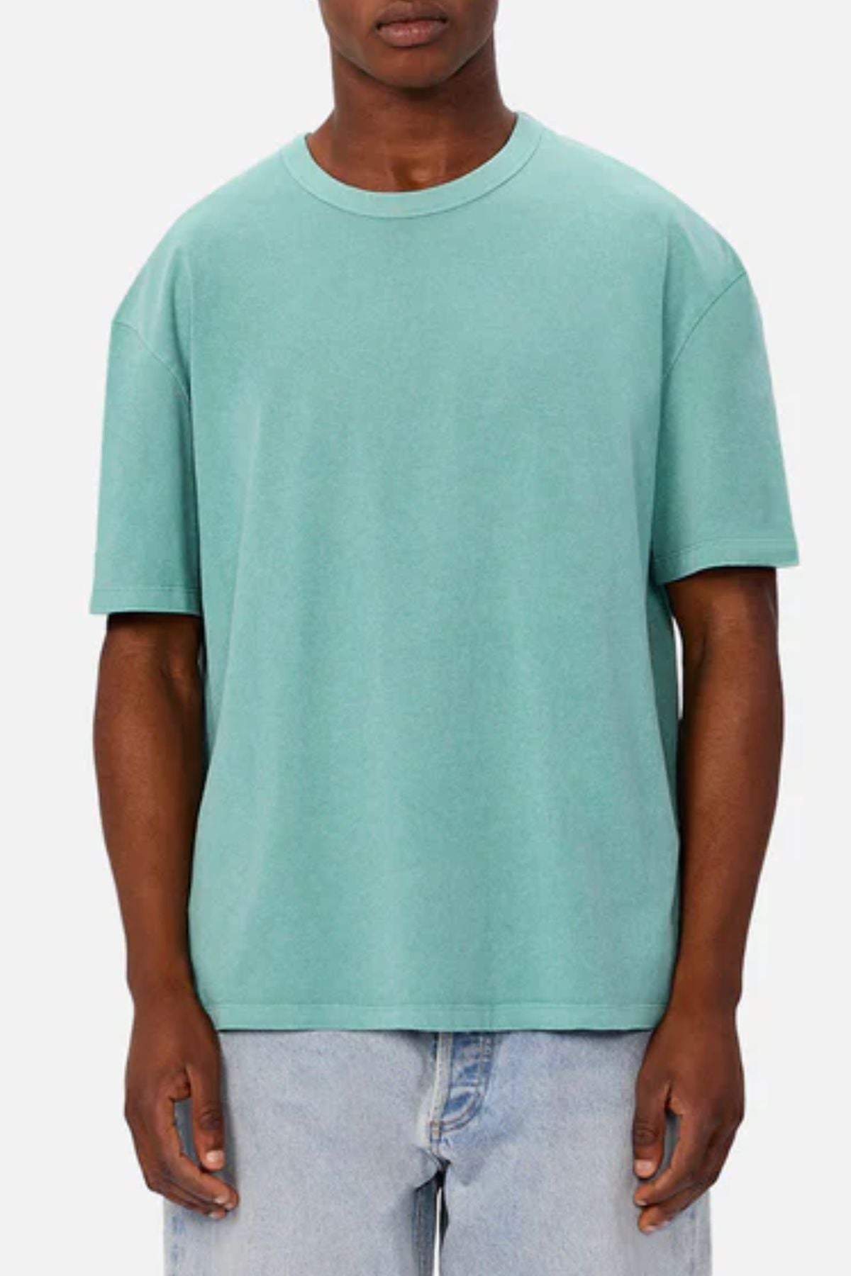 A person in the Industrie The Del Sur Tee, a loose-fitting short-sleeve light teal oversized t-shirt, paired with light blue jeans stands facing forward against a plain gray background. Their face is partially obscured.