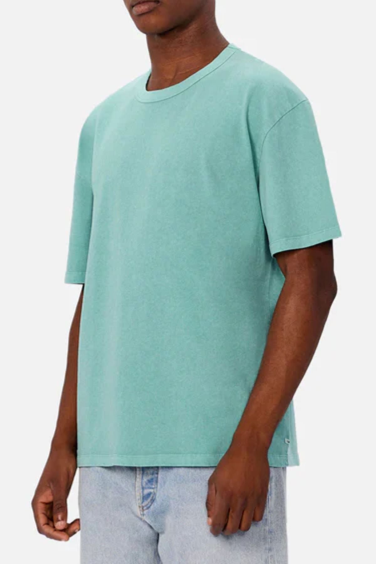 A person in an oversized Industrie The Del Sur Tee in plain teal and light blue jeans stands before a light grey backdrop. The relaxed fit and partially out-of-frame head create a modern, minimal style.