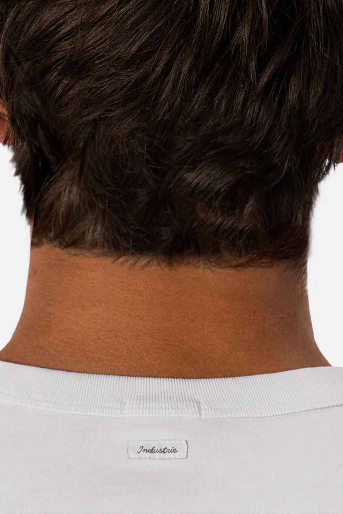 The image features the back of a person's head and neck with short brown hair. They are wearing a white, comfortable tee called "Industrie The Del Sur Tee," made from 100% cotton, with a small label displaying the word "Industrie.