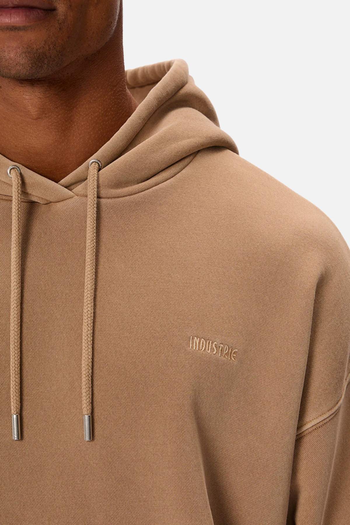 A person in an oversized beige Industrie The Del Sur Washed Hoodie with drawstrings, featuring "INDUSTRIE" on the chest. Cropped to show only the upper torso and part of the face against a plain white background, it exudes a relaxed vibe.