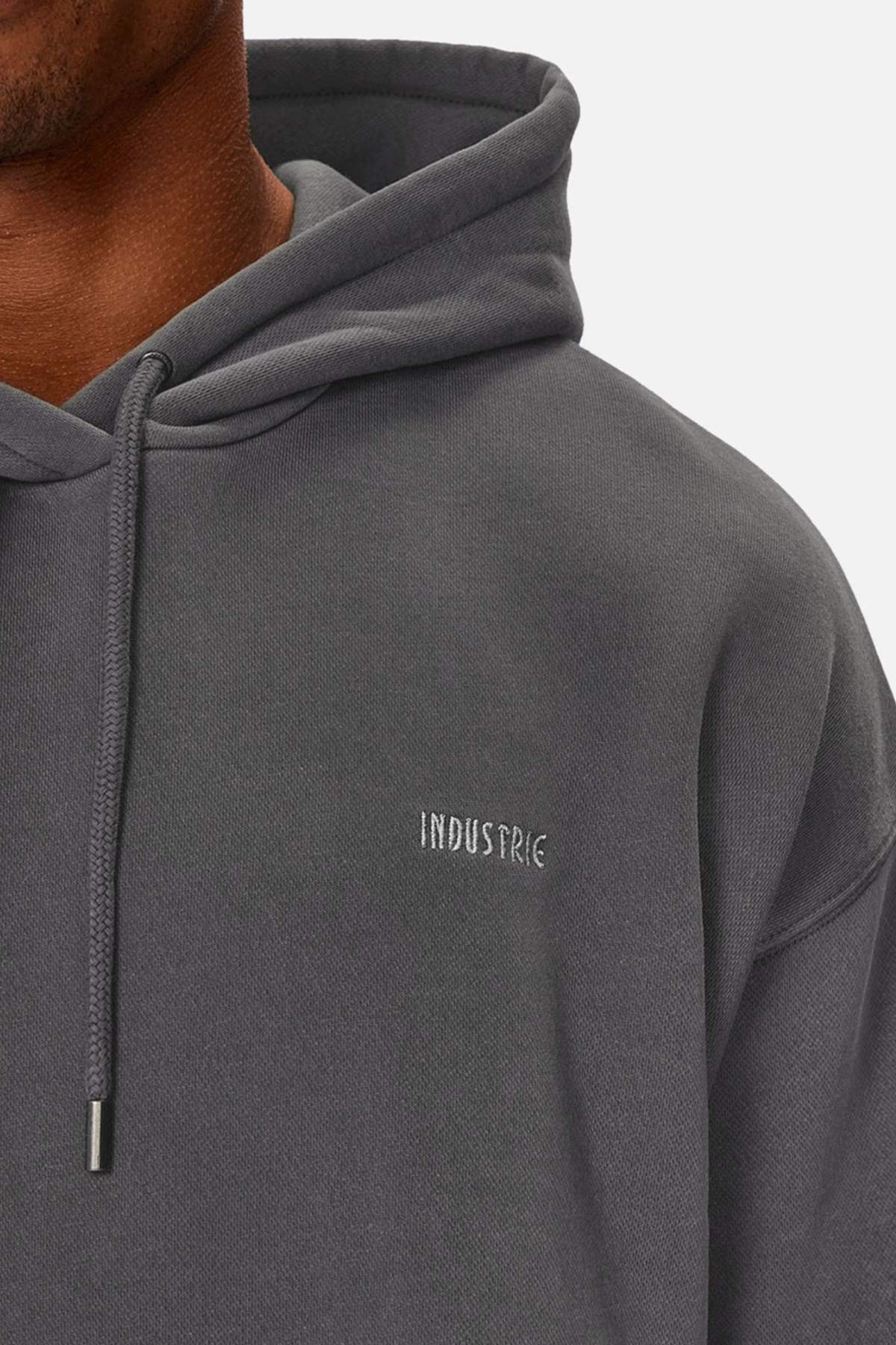 Close-up of a person wearing the Industrie The Del Sur Washed Hoodie in dark gray acid wash, featuring “INDUSTRIE” embroidered in small white letters on the chest. Only the lower face and upper body are shown, emphasizing its oversized fit.